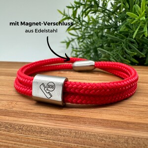 Könnte beinhalten: Rotes Paracord-Armband mit einem magnetischen Verschluss aus Edelstahl und einem silbernen Anhänger mit einem Telefon und dem Text "SOS".
