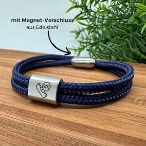 Könnte beinhalten: Ein marineblaues Seilarmband mit einem Magnetverschluss aus Edelstahl. Das Armband hat einen rechteckigen Verschluss mit einem SOS-Telefon-Symbol. Der Text "mit Magnet-Verschluss aus Edelstahl" befindet sich oben links.