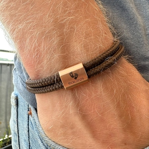 Può includere: Un braccialetto in corda marrone con un ciondolo a barra in oro rosa inciso con un simbolo di impronta e la data "15.06.2024".