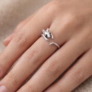 Pu&ograve; includere: Un anello color argento a forma di topo, con occhi neri e una coda ricurva. Le braccia del topo si avvolgono attorno al dito, creando un design aperto. L'anello &egrave; indossato su un dito.