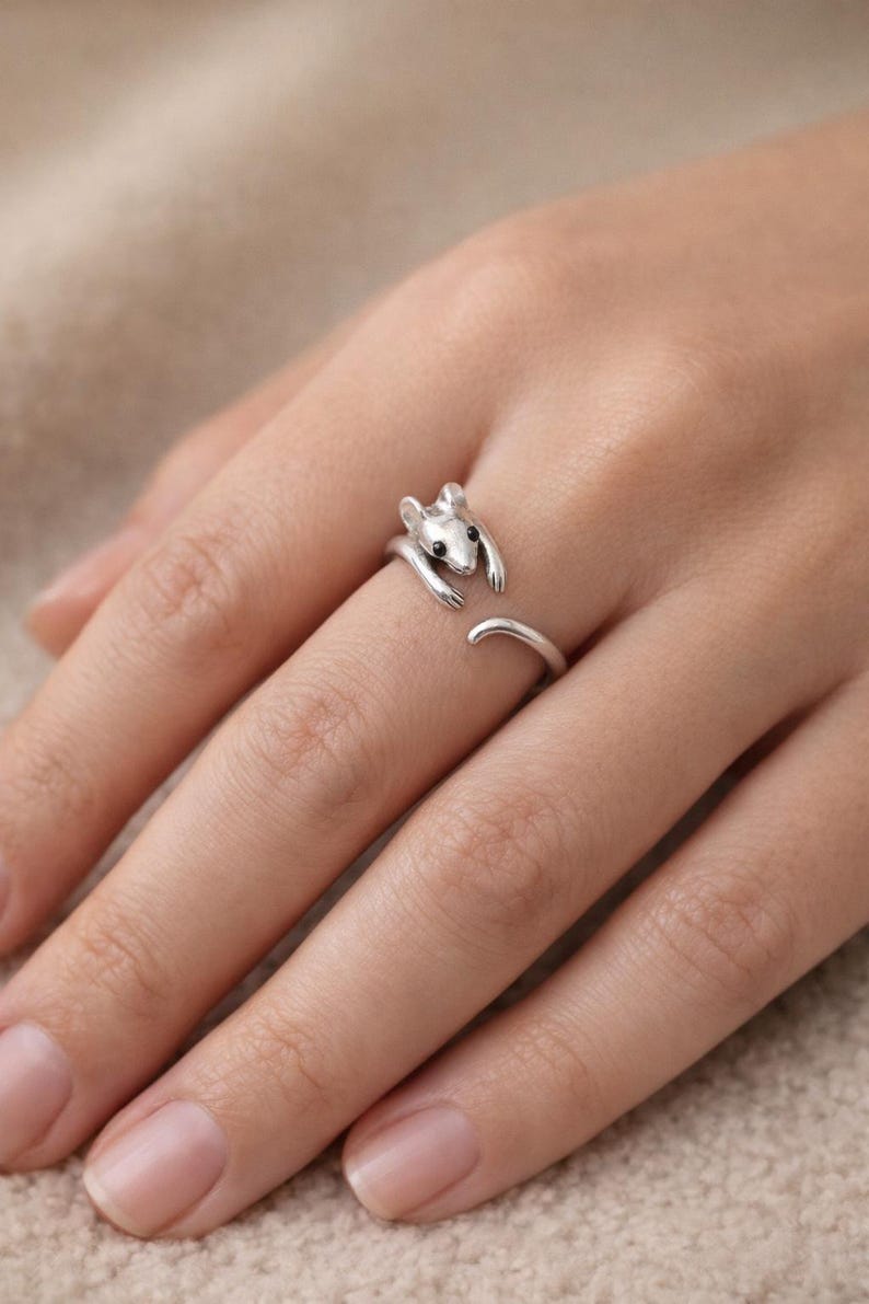 Pu&ograve; includere: Un anello color argento a forma di topo, con occhi neri e una coda ricurva. Le braccia del topo si avvolgono attorno al dito, creando un design aperto. L'anello &egrave; indossato su un dito.