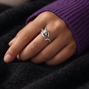 Pu&ograve; includere: Un anello color argento a forma di scoiattolo. L'anello presenta un design a scoiattolo, con la testa e le zampe che avvolgono il dito e la coda che si estende lungo il lato. L'anello &egrave; indossato su una mano, con un maglione viola e una coperta nera sullo sfondo.