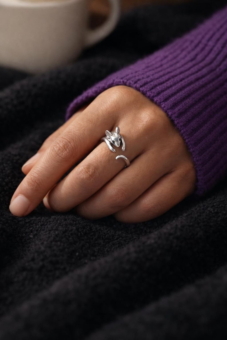 Pu&ograve; includere: Un anello color argento a forma di scoiattolo. L'anello presenta un design a scoiattolo, con la testa e le zampe che avvolgono il dito e la coda che si estende lungo il lato. L'anello &egrave; indossato su una mano, con un maglione viola e una coperta nera sullo sfondo.