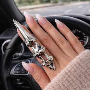 Op de afbeelding: Zilverkleurige vingerring met een klauwachtig ontwerp. De ring heeft een lange, puntige punt en een gedetailleerde, gelede structuur. De ring wordt gedragen op een hand met lange, ovaalvormige nagels die lichtroze zijn geverfd.