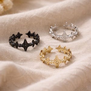May include: Three open-ended rings with star designs in black, silver, and gold. The rings are made of metal and rest on a beige fabric surface. The star designs are evenly spaced around the bands.