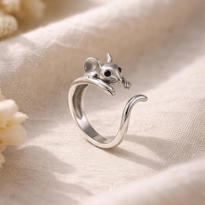 May include: A silver-colored ring featuring a mouse design. The mouse has black eyes and is perched on the ring's band. The ring has a curved tail design. The ring is likely made of sterling silver or a similar metal.