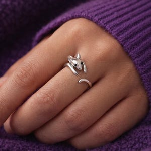 Pu&ograve; includere: Un anello color argento a forma di topo, con la testa e le braccia del topo che avvolgono il dito. L'anello ha occhi neri e una coda ricurva. L'anello &egrave; mostrato su una mano, su uno sfondo viola.