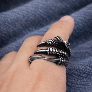 Könnte beinhalten: Ein silberfarbener Krallenring mit scharfen, spitzen Krallen. Der Ring umschließt den Finger und erzeugt einen wilden und kantigen Look. Der Ring hat strukturierte Schuppen und ein dunkles Finish.