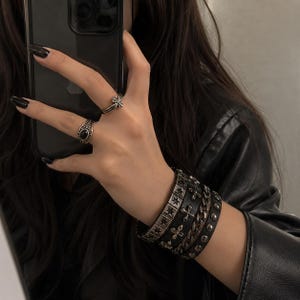 May include: A set of four stacked bracelets featuring black leather bands with silver-toned metal star, cross, chain, and stud embellishments, paired with two silver-toned rings, one featuring a black oval stone and the other a cross design.