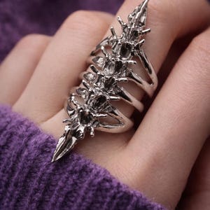 May include: A silver-colored, articulated ring with a spine-like design. The ring features multiple bands and a central, pointed element, creating a gothic or fantasy aesthetic. The ring is worn on a finger, against a purple background.