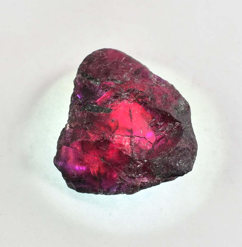 May include: A raw, natural gemstone in a deep burgundy hue, with a bright, glowing interior. The stone has a textured surface with dark, irregular edges, and a bright, reflective core. The gemstone is set against a white background.