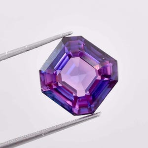 Puede incluir: Una gema facetada de corte cuadrado, de color púrpura intenso, sujeta con pinzas plateadas. Las facetas de la gema reflejan la luz, mostrando tonos de violeta y azul. Una pieza de joyería.