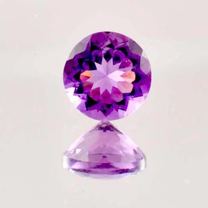 May include: A round, faceted amethyst gemstone in shades of purple. The gem's facets catch the light, creating a sparkling effect. The base of the gem is also visible, creating a mirrored reflection. The background is a light, reflective surface.