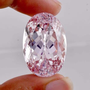 May include: A large, oval-cut pink gemstone with a faceted surface. The gem is held between fingers, showcasing its light pink hue and intricate internal reflections. The gemstone's shape and cut create a brilliant display of light.
