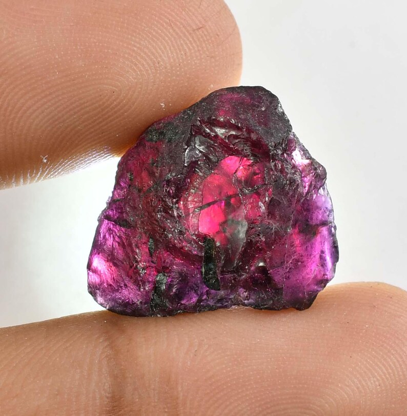 May include: A raw, natural gemstone in a deep magenta colour. The gem has a crystalline structure with dark, almost black, inclusions. The stone is held between two fingers, showing its irregular shape and reflective surfaces.