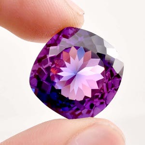 Peut inclure: Une grosse pierre précieuse taillée en coussin, d'un violet éclatant. La gemme présente des facettes complexes qui créent un motif en étoile au centre, reflétant la lumière. La pierre est tenue entre deux doigts sur fond blanc.