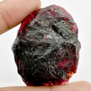 Puede incluir: Una gema en bruto, sin cortar, de color rojo intenso, que presenta una superficie rugosa y texturizada. La piedra tiene zonas de coloración oscura, casi negra, que contrastan con el rojo vibrante. La forma general es irregular.