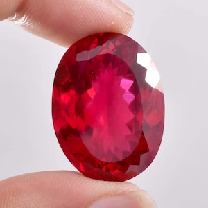 Puede incluir: Una gran gema de rubí facetada de forma ovalada. La gema es de un color rojo intenso y vibrante con múltiples facetas que reflejan la luz. El rubí se sostiene entre dos dedos sobre un fondo blanco.