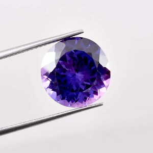 Può includere: Una gemma rotonda e sfaccettata con una tonalità viola intenso, tenuta da pinzette d'argento. Le sfaccettature della gemma riflettono la luce, creando una brillante esposizione di colore. La gemma è il punto focale su sfondo bianco.