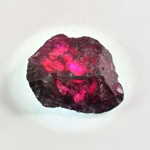 May include: A natural garnet gemstone, uncut and rough. The stone is a deep red with bright pink and purple highlights. The surface has a textured, crystalline appearance, with a dark, almost black, outer layer.