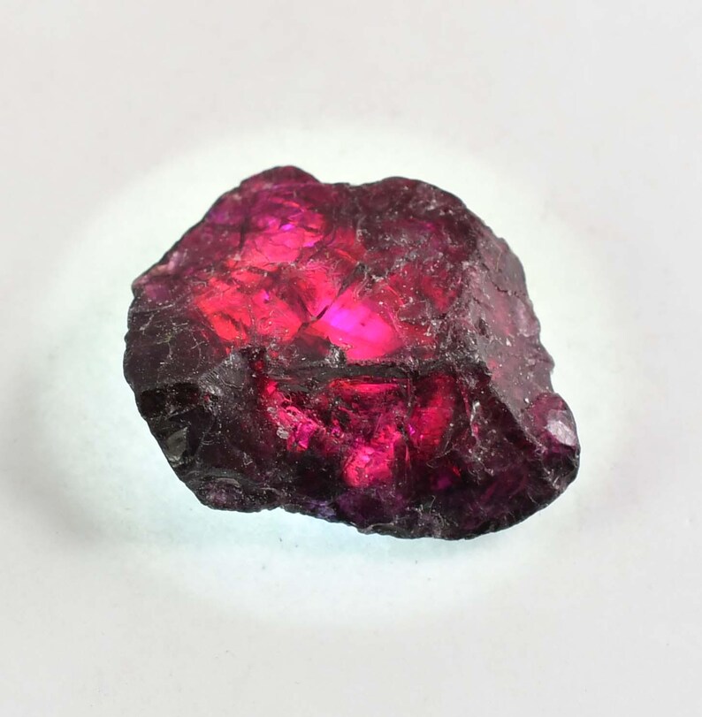 May include: A natural garnet gemstone, uncut and rough. The stone is a deep red with bright pink and purple highlights. The surface has a textured, crystalline appearance, with a dark, almost black, outer layer.
