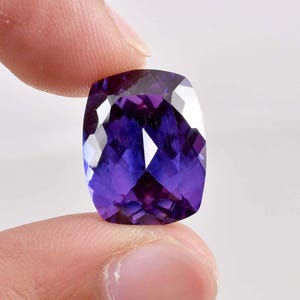 Puede incluir: Una gema facetada, de talla cojín, con tonos morados y azules profundos. La gema se sostiene entre dos dedos, mostrando sus superficies pulidas y reflejos internos. El color de la piedra transita del morado oscuro a un azul vibrante.