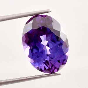 Puede incluir: Una gema de tanzanita ovalada y facetada, sujeta con pinzas. La gema presenta un color azul-violeta intenso y múltiples facetas que reflejan la luz. El fondo es blanco. Un ejemplo de la gema.