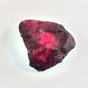 May include: A natural ruby gemstone, rough-cut with a deep red colour and a slightly translucent appearance. The stone has a jagged, uneven surface and a dark, almost black, outer layer.