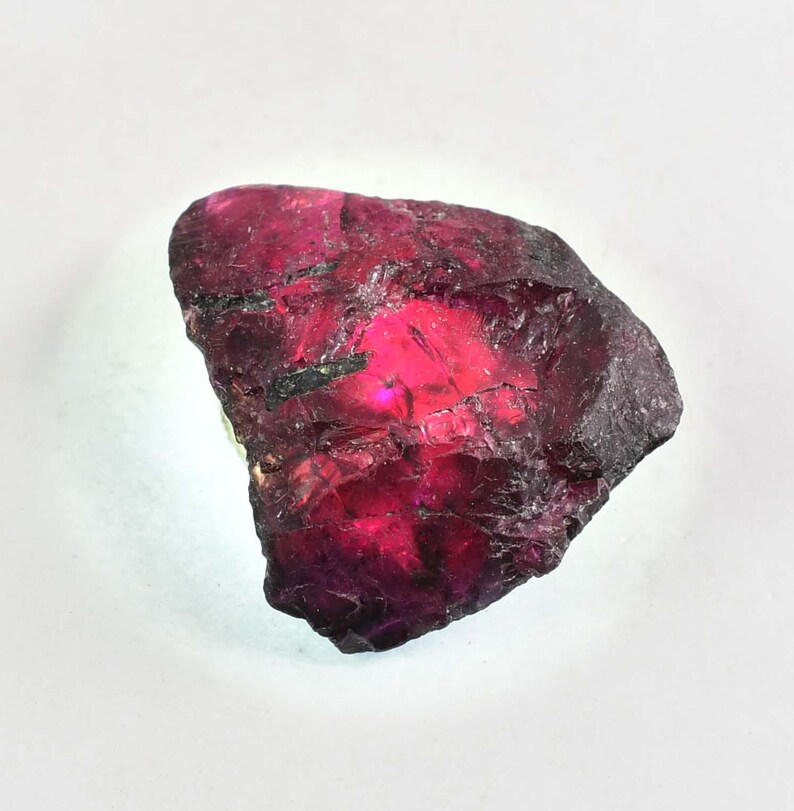 May include: A natural ruby gemstone, rough-cut with a deep red colour and a slightly translucent appearance. The stone has a jagged, uneven surface and a dark, almost black, outer layer.