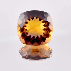 May include: A faceted, cushion-cut gemstone with a starburst design in the center. The gem features a gradient of colors, transitioning from a deep brown at the top to a vibrant yellow-orange in the center. The gem is reflected below.