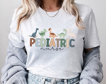 May include: A white t-shirt with a graphic of a line of colorful dinosaurs and the text "PEDIATRIC nurse".