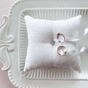 Può includere: Un cuscino portafedi in lino bianco con due fedi nuziali d'oro legate con un nastro di raso bianco.