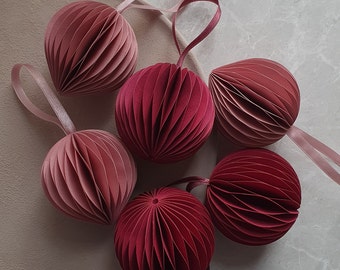 May include: Six paper ornaments in shades of pink and red. The ornaments are folded into a geometric pattern and have a textured surface.