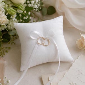 May include: A white ring pillow with two gold wedding bands. The pillow is square and has a satin ribbon tied in a bow. The rings are held in place by the ribbon. The background includes flowers and a card.