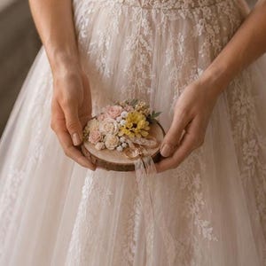 May include: A circular wooden slice ring bearer pillow decorated with an arrangement of small artificial pink, yellow, and white flowers. A thin, light-colored ribbon is attached to the wood, looped to hold two gold-toned metal rings securely.