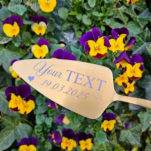 Op de afbeelding: Gouden taartschep met de tekst "Your TEXT 19.03.2025" en twee kleine harten gegraveerd op de voorkant. De schep is omringd door paarse en gele viooltjes.
