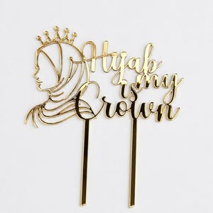 Op de afbeelding: Een goudkleurige taarttopper met de tekst "Hijab is my Crown" in sierlijke letters. Het ontwerp bevat een silhouet van een vrouw met een hijab en een kroon. De topper heeft twee lange, dunne stelen om in een taart te steken.