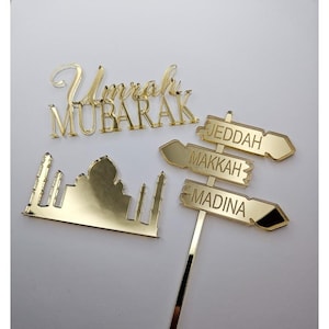 Op de afbeelding: Gouden spiegelende acryl taarttopper met de woorden "Umrah Mubarak" en een silhouet van een moskee. Drie gouden spiegelende acryl borden met pijlen wijzen naar "Jeddah", "Makkah" en "Madina".