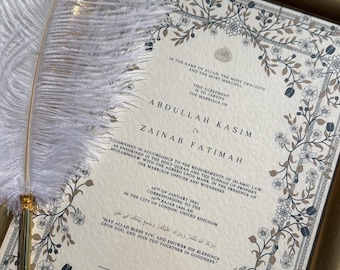 May include: A framed wedding certificate with floral border and text. The names Abdullah Kasim and Zainab Fatimah are featured. A white feather pen rests on the frame. The certificate is on a white satin surface.