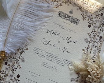 Puede incluir: Un certificado de matrimonio con borde floral y texto en caligrafía árabe. El documento incluye los nombres Abdel Majeed, Sarah & Noora. Una pluma blanca y un ramo de flores secas se colocan cerca del certificado.