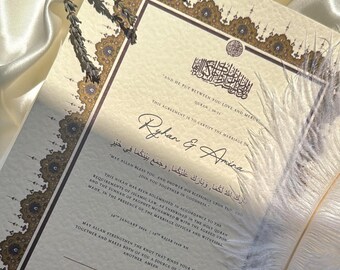 May include: A marriage certificate with gold and brown decorative borders, featuring Arabic calligraphy and English text. A white feather quill and dried lavender sprigs are placed on the document, against a satin background.