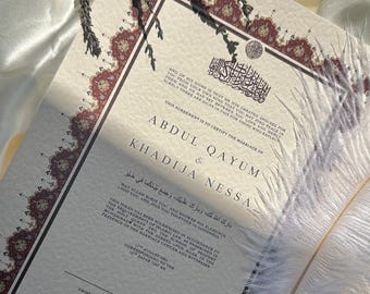 Puede incluir: Un certificado de boda con detalles en burdeos y dorado, con los nombres ABDUL QAYUM & KHADIJA NESSA. Una pluma blanca descansa junto al documento, con ramitas de lavanda seca. El certificado está sobre una superficie de satén blanco.