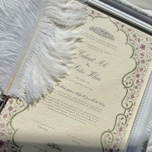 May include: A framed wedding certificate with floral accents and calligraphy. A white feather quill pen rests on the document. The certificate includes the names Cahmid Ali and Sadia Khan, and the date 18.08.25.