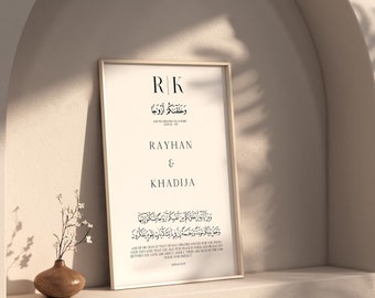 Peut inclure: Une impression encadrée avec un fond blanc et du texte noir. Le texte comprend les initiales "R|K", les noms "Rayhan & Khadija" et une inscription arabe. L'impression est sur une étagère dans une pièce avec un mur blanc et une arche incurvée.