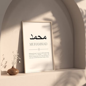 May include: A framed print with the Arabic word for Muhammad in black calligraphy. The word "Muhammad" is written in English below the Arabic text. The print has a white background and a thin black frame. The print is sitting on a shelf.