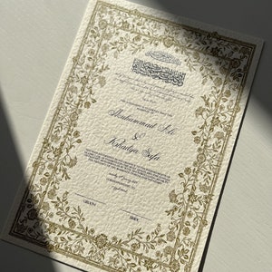 May include: A gold-coloured wedding certificate with ornate floral borders. The text reads "This agreement is to certify that the marriage of Muhammad Ali & Khadija Safa has been solemnised in accordance to the requirements of Islamic Law, as enshrined in the Holy Quran and the Sunnah of Prophet Muhammad (PBUH) with the agreed upon mahr, in the presence of the marriage officer and witnesses corresponding to Groom Bride."