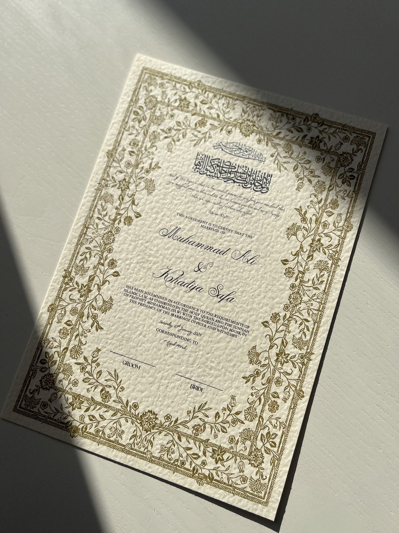 May include: A gold-coloured wedding certificate with ornate floral borders. The text reads "This agreement is to certify that the marriage of Muhammad Ali & Khadija Safa has been solemnised in accordance to the requirements of Islamic Law, as enshrined in the Holy Quran and the Sunnah of Prophet Muhammad (PBUH) with the agreed upon mahr, in the presence of the marriage officer and witnesses corresponding to Groom Bride."