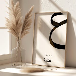May include: A framed print with a black calligraphy design of the Arabic word "Inaya" which means "grace" in English. The print is on a white background with a gold frame. The name "Inaya" is written in English below the calligraphy.