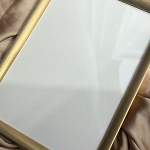 Puede incluir: Un marco de fotos de color dorado con un paspart&uacute; blanco. El marco est&aacute; sobre una superficie de tela beige.