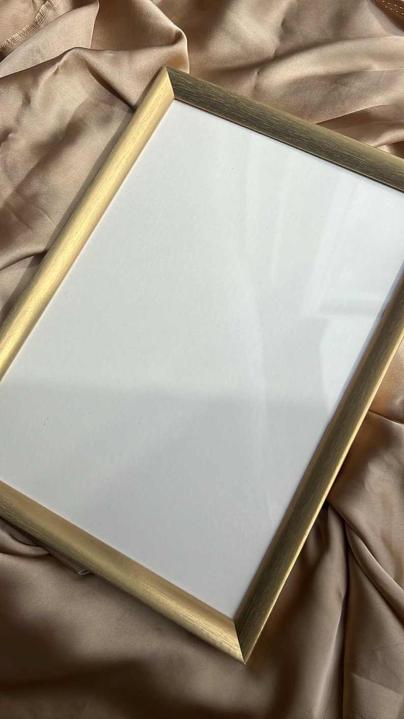 Puede incluir: Un marco de fotos de color dorado con un paspart&uacute; blanco. El marco est&aacute; sobre una superficie de tela beige.